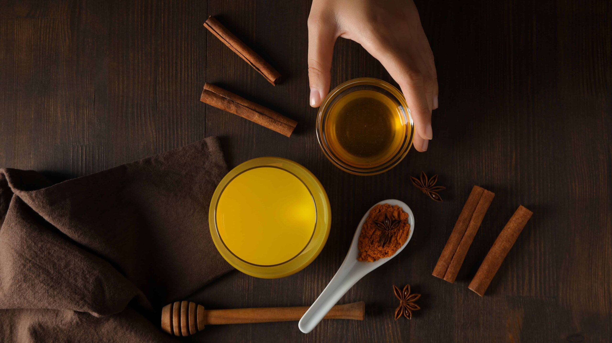 Top-down view of a dark wooden table with cinnamon sticks, star anise, a jar of honey, and a white spoon filled with ground cinnamon next to a yellow-lidded jar.